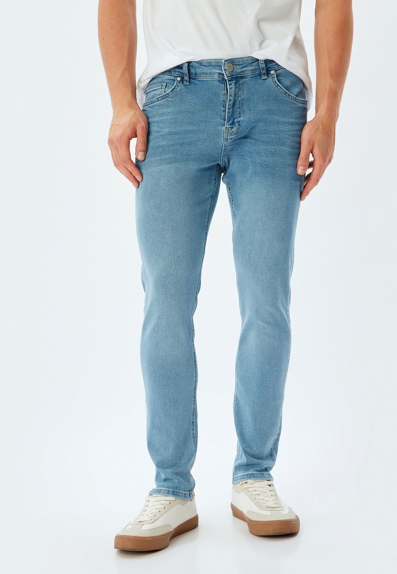 Jeans de mezclilla azul claro con un diseño ajustado, que cuentan con cinco bolsillos, pasadores para cinturón y un cierre de botón. Combinados con zapatillas blancas y marrones claras.