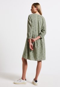 Abito in cotone verde con ricami ad occhio, maniche tre quarti, vita arricciata e gonna a ruota. Indossato con sneaker bianche.