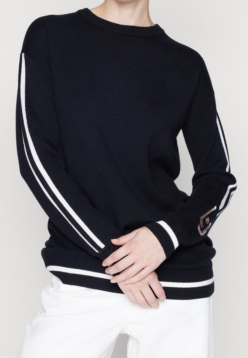 Personne portant un pull noir avec des rayures blanches sur les manches et l'ourlet, détail brodé sur le poignet, associé à un pantalon blanc.
