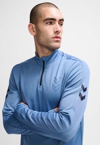 Lichtblauw sporttop met kwartrits, gemaakt van gestructureerde stof, met zwarte chevronpatronen op de mouwen en een subtiel logo op de borst.