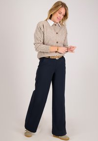 Lichtbruin gebreide trui met ronde kraag en knopen, gecombineerd met marineblauwe wijde broek met een riem van tan leer. Schoenen zijn lichtgrijs.