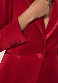Rød fløjl blazer med snipperkrave, enkelt knaplukning og fire matchende knapper på manchetterne. Glat tekstur med struktureret design.