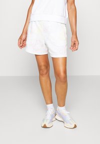 Osoba oblečená v šortkách s bílým mramorovým vzorem, bílých ponožkách, tenisových botách v pastelových barvách a bílém topu bez rukávů na jednobarevném pozadí.