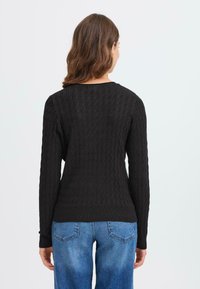 Svart strikkegenser med en strukturert kabelmønster, lange ermer og rund hals. Hemmen er litt ribbet. Brukt sammen med blå denimjeans.