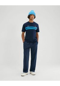Camiseta azul marino con rayas azul claro en el pecho, combinada con pantalones azul marino y zapatillas blancas. El modelo lleva un gorro de punto azul claro.