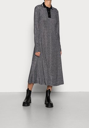 Kobieta w długim rękawem, błyszczącej szarej sukience midi z czarnym kołnierzykiem i czarnymi botkami do kostek, stojąca na tle białego, jednolitego tła.