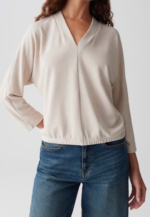 Femme portant un haut beige côtelé à col en V et manches longues, ainsi qu'un jean bleu, debout avec une main sur la hanche.