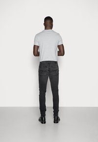 En man står vänd mot en vägg iklädd en ljusgrå t-shirt, mörka slim-fit jeans och svarta läderstövlar på ett grått golv.