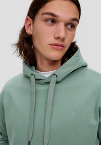Groene hoodie met een trekkoordcapuchon, gemaakt van zachte stof. Heeft een klein geborduurd logo "QS" op de borst en een gladde textuur.