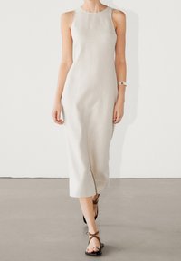 Robe en lin beige clair avec un design sans manches, un col rond et une fente latérale. Associée à des sandales à lanières marron.