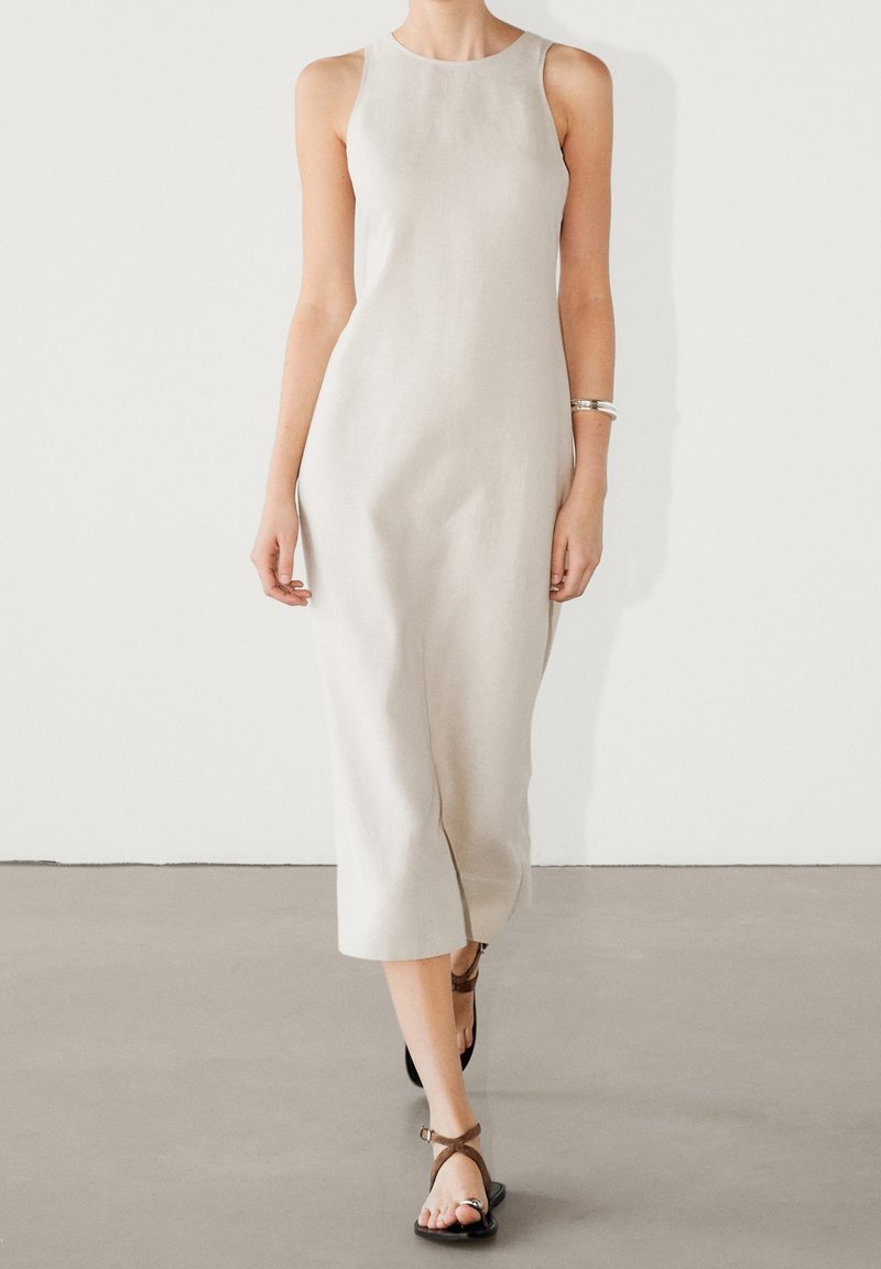 Robe en lin beige clair avec un design sans manches, un col rond et une fente latérale. Associée à des sandales à lanières marron.