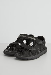 Svarta sandaler med justerbara remmar, texturerade sulor och utstuket mönster. Förstärkt häl och tå, märkt med "Timberland"-logotyp.