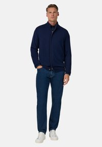 Marineblauer Reißverschluss-Pullover aus weichem Material, kombiniert mit blauen Jeans und weißen Sneakers. Das Outfit zeichnet sich durch ein minimalistisches Design und eine legere Passform aus.