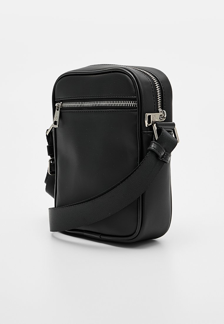 Bolso negro de bandolera hecho de material sintético, con una textura suave, herrajes plateados, un bolsillo con cremallera y una correa ajustable.