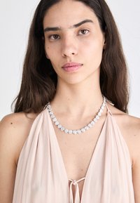 Collar de plata en forma de corazón con piedras brillantes, llevado sobre una prenda drapeada de color rosa claro. Diseño simple con un cierre seguro.