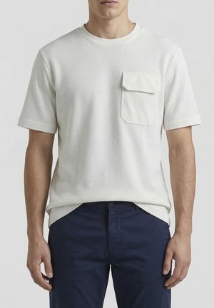 Muškarac odjeven u bijelu teksturiranu košulju s kratkim rukavima i preklopnim džepom na prsima te tamnoplave hlače, stoji ispred jednobojne pozadine.