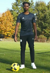 Zwart sportshirt met Adidas-logo, zwarte broek met embleem, witte voetbalschoenen. Gele en zwarte voetbals met op een grasveld, herfstbomen op de achtergrond.