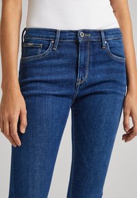 Sötétkék farmernadrág, slim fit szabással, öt zsebbel, látható elülső gombbal és narancssárga varrással. Sima felület.