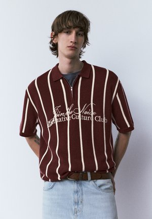 Junger Mann mit braun gestreiftem kurzärmeligem Poloshirt mit Text, hellblauer Jeans, steht vor einem schlichten hellen Hintergrund.