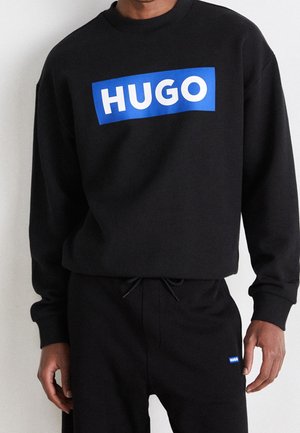 Czarna bluza i spodnie z niebieskimi prostokątnymi logo "HUGO" noszone przez osobę pokazana od ramion do bioder.