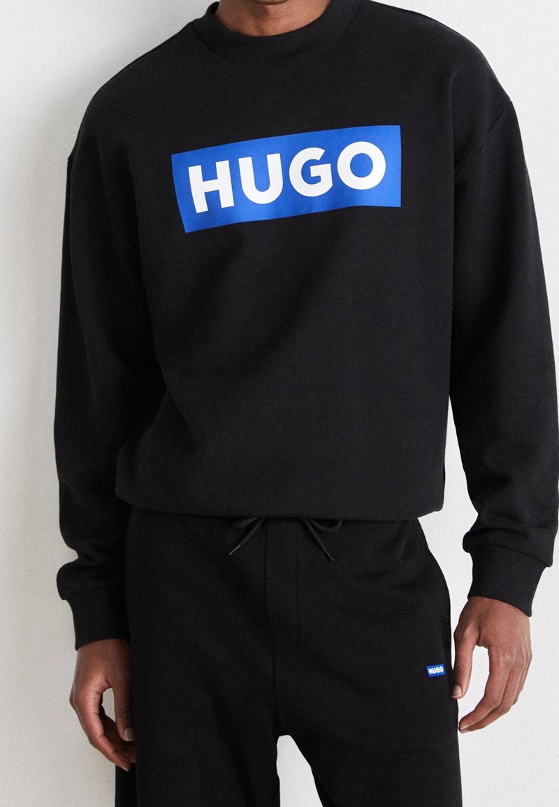 Sweat-shirt noir et pantalon avec des logos rectangulaires bleus "HUGO" portés par une personne montrée des épaules aux hanches.