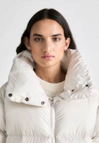 Puhasta jakna v svetlem bež odtenku z visokim, strukturiranim ovratnikom. Ima zadrgo in vidno šivanje z dodatki na gumbih.