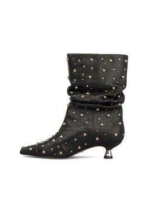 Bota negra de piel hasta media pantorrilla con caña caída, punta afilada, tacón bajo metálico en bloque y adornos con tachuelas doradas dispersas.