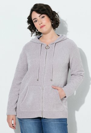 Ung kvinde med kort krøllet hår iført en lysegrå lynlåshoodie og blå jeans med den ene hånd i lommen.