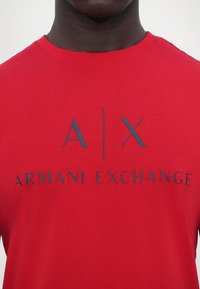 Raudona medvilninė marškinė su trumpomis rankovėmis, featuring tamsiai pilką logotipą: "A|X ARMANI EXCHANGE", atspausdintą per krūtinę. Lygus tekstūra.