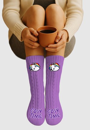 Person sitzt mit angewinkelten Knien, hält eine braune Tasse und trägt lila Socken mit Einhornköpfen und dem Schriftzug "COSY TIME" auf den Füßen.