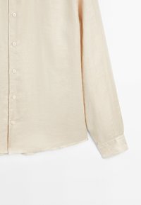 Camicia lunga beige chiaro con maniche, realizzata in tessuto morbido e testurizzato, con bottoni frontali e orlo arrotondato. Presenta bottoni abbinati e maniche a polsino.