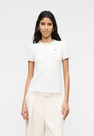 Žena s dlhými tmavými vlasmi, oblečená v bielom priliehavom tričku s krátkym rukávom a zeleným lemom a v svetlo béžových nohaviciach, stojaca pred jednoduchým pozadím.