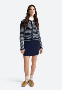 Cardigan lavorato a maglia a righe blu e bianche con bottoni tondi blu, abbinato a una gonna blu, calze crema e scarpe marroni.