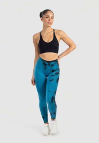 Svart sport-bh med veckad detalj tillsammans med blå och svart tie-dye leggings. Modellen står med händerna på höfterna och bär vita sneakers.