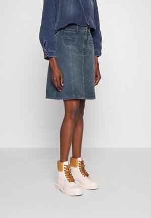 Kvinde iført en falmet blå denimnederdel, mørkeblå langærmet top og cremefarvede snørestøvler med lysebrune polstrede kraver.