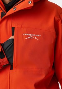 Giacca impermeabile rossa con tasca con zip e dettagli neri. Presenta il logo "SWEDEMOUNT" in bianco sopra un grafico di montagna. Tessuto strutturato.