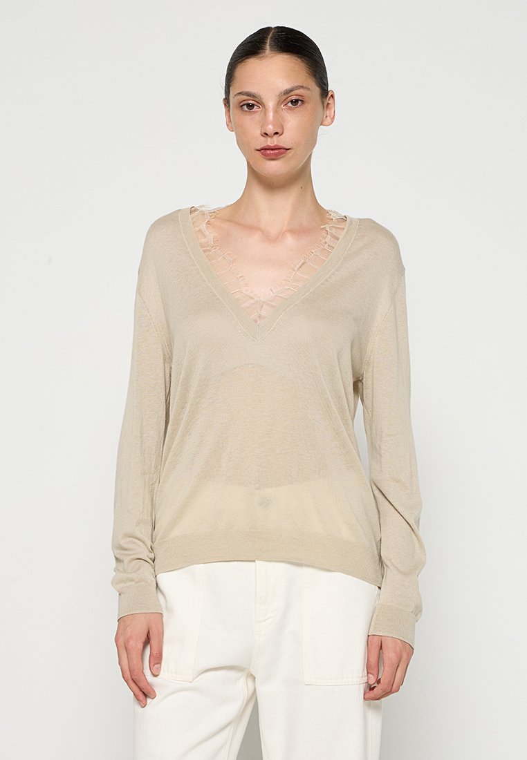 Vrouw in een beige V-hals trui met doorschijnende ruches en witte broek, staande tegen een effen lichte achtergrond.