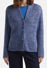 Frau, die eine blaue Strickjacke mit drei Knöpfen über einem blauen Hemd und einer dunklen Hose trägt, steht mit entspannten Händen an den Seiten.