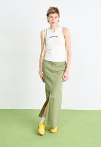 Haut sans manches blanc avec l'imprimé "DICKIES", associé à une jupe maxi verte avec une fente sur le côté et des baskets jaunes. Textures de vêtements naturelles.