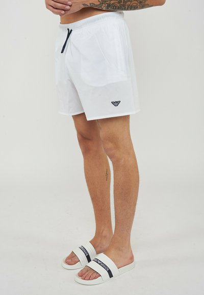 Homme portant un short de bain blanc avec un logo noir et des sandales slides blanches Armani, debout devant un fond uni.