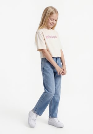 Ξανθιά κοπέλα που χαμογελά, φοράει μια μπεζ κοντομάνικη μπλούζα με ροζ γράμματα, ανοιχτόχρωμο μπλε τζιν και λευκά αθλητικά παπούτσια, στέκεται στο ένα πόδι.