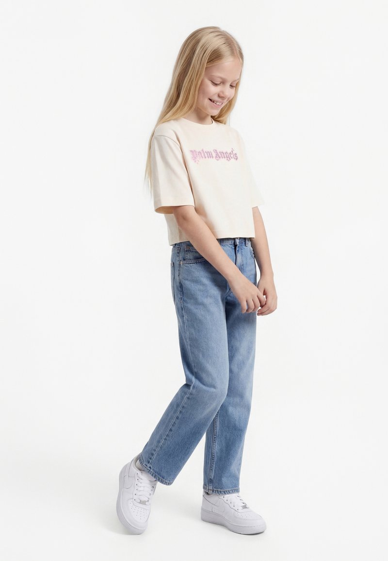 Blondynka uśmiechająca się, ubrana w beżowy krótki t-shirt z różowym napisem, jasnoniebieskie dżinsy i białe trampki, stojąca na jednej nodze.