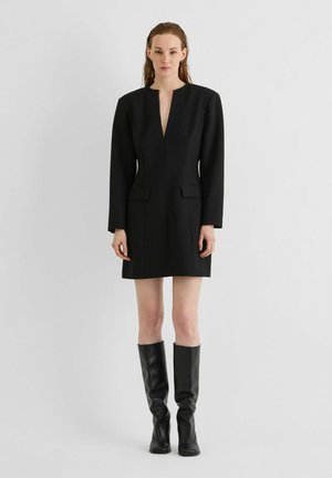 Femme debout portant une robe blazer noire à manches longues avec poches à rabat et bottes noires montantes sur un fond clair uni.