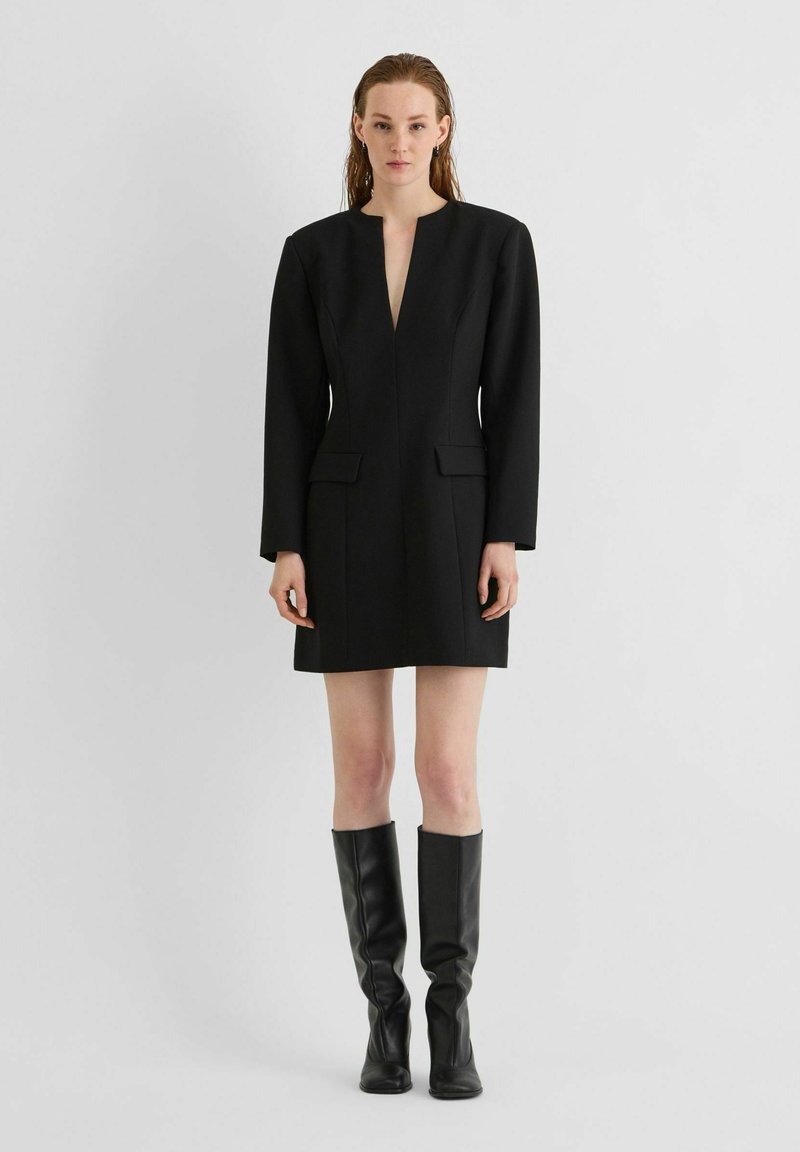 Femme debout portant une robe blazer noire à manches longues avec poches à rabat et bottes noires montantes sur un fond clair uni.