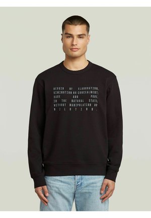 Homme portant un sweat-shirt noir avec du texte blanc et un jean bleu clair, debout devant un fond uni clair.