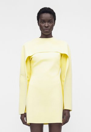 Kobieta ubrana w bladżółtą mini sukienkę z długim rękawem i strukturalnym, pelerynopodobnym detalem na ramionach, stojąca na jednolitym tle.