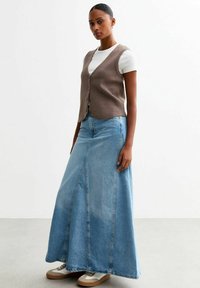 Chaleco de punto marrón sobre una camiseta blanca, combinado con una falda larga de mezclilla azul claro y zapatillas beige, con un corte acampanado y costuras laterales.