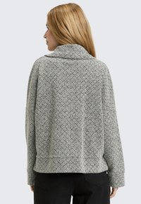 Vrouw met lang blond haar, gekleed in een gestructureerde lichtgrijze trui en zwarte broek, gezien van achteren tegen een effen achtergrond.