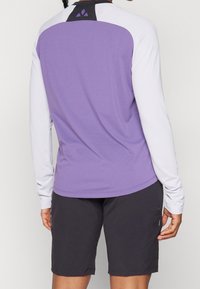 Tröja med långa ärmar i athletic-stil, med ett lila nätmönster och vita ärmar; har en svart detalj vid halsringningen. Matchas med mörka shorts.