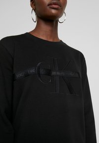 Musta puuvillapusero, jossa on brodeerattu "CK"-logo ja musta nauha, jossa lukee "CALVIN KLEIN JEANS". Pyöreä pääntie ja ribbaatut hihansuut.